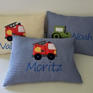 Könnte beinhalten: Drei blaue, cremefarbene und beigefarbene Waffel-Kissen mit gestickten Feuerwehrautos und Traktoren. Die Kissen haben die Namen "Val", "Moritz" und "Noah" gestickt.