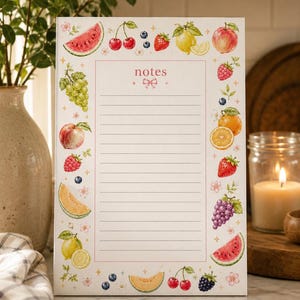 May include: A rectangular notepad with a white background and a central lined section for writing. The word notes is printed at the top with a small pink bow. The border features watercolour illustrations of various fruits including watermelon, cherries, grapes, peaches, raspberries, lemons, oranges, and blackberries.