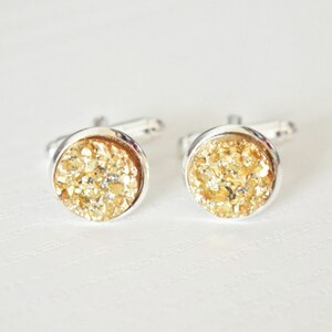 May include: A pair of round cufflinks with a silver-colored metal setting. The center of each cufflink is filled with a sparkling, golden-colored material. The cufflinks are displayed on a white surface.