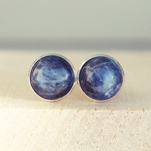 May include: A pair of round stud earrings with a silver bezel. Each earring features a blue gemstone with white marbling. The earrings are displayed on a light-colored wooden surface.