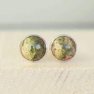 May include: A pair of round stud earrings featuring a natural stone with green and pink hues. The stones are set in a silver-colored bezel. The earrings are displayed on a light-colored wooden surface.