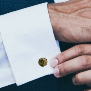 May include: Close-up of a person's hands adjusting a white dress shirt cuff with a gold-toned cufflink. The cufflink has a circular design with a dark, textured appearance. The person is wearing a navy blue vest.