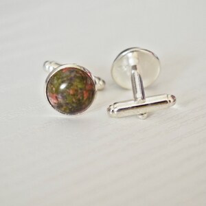 May include: A pair of cufflinks with a silver finish. Each cufflink features a round, polished stone with a mix of green, brown, and red hues. The cufflinks are displayed on a white surface.
