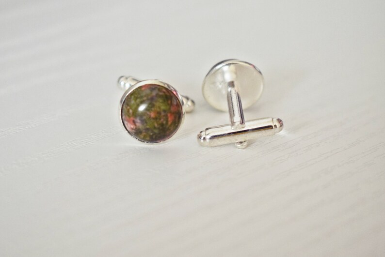 May include: A pair of cufflinks with a silver finish. Each cufflink features a round, polished stone with a mix of green, brown, and red hues. The cufflinks are displayed on a white surface.
