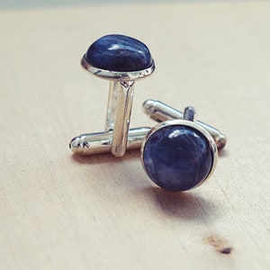 May include: A pair of silver cufflinks with a dark blue, rounded stone set in each cufflink. The cufflinks are displayed on a light-colored wooden surface.
