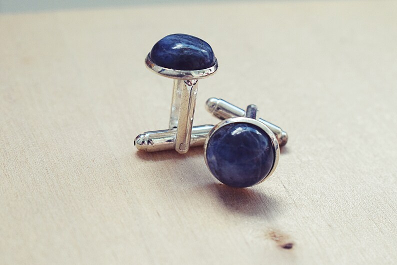 May include: A pair of silver cufflinks with a dark blue, rounded stone set in each cufflink. The cufflinks are displayed on a light-colored wooden surface.
