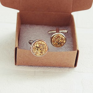 May include: A pair of gold-colored cufflinks with a textured, glittery surface, presented in a small, open, brown cardboard box. The cufflinks have a silver-colored metal border and are set against a white background.