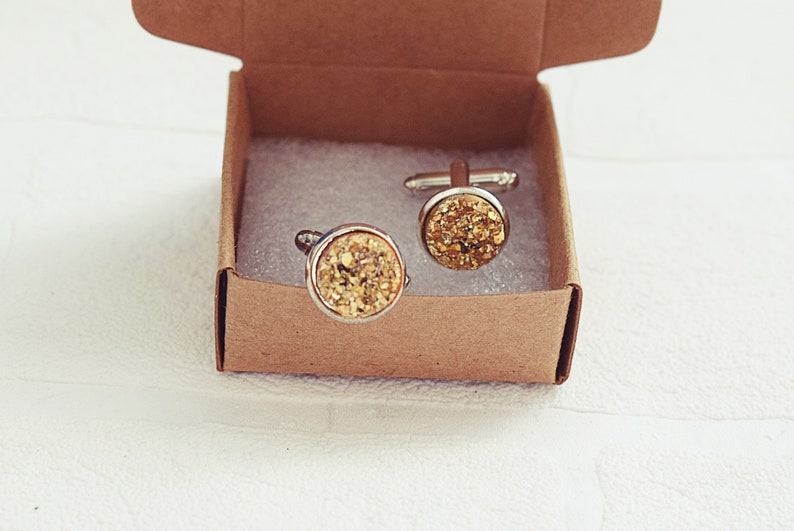 May include: A pair of gold-colored cufflinks with a textured, glittery surface, presented in a small, open, brown cardboard box. The cufflinks have a silver-colored metal border and are set against a white background.