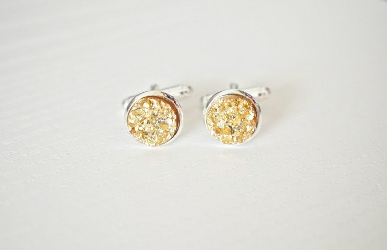May include: A pair of round cufflinks with a silver-colored metal setting. The center of each cufflink is filled with a sparkling, golden-colored material. The cufflinks are displayed on a white surface.