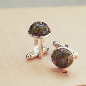 May include: A pair of silver cufflinks with round, polished unakite stone faces. The unakite stones display a mix of green and pink hues. The cufflinks are set against a light wooden surface.