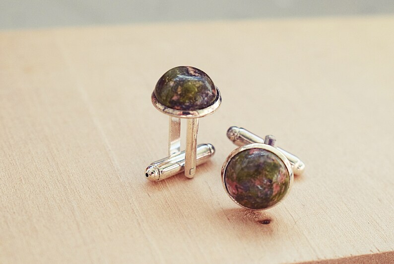 May include: A pair of silver cufflinks with round, polished unakite stone faces. The unakite stones display a mix of green and pink hues. The cufflinks are set against a light wooden surface.
