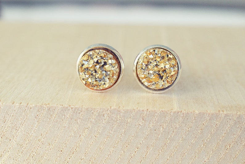 May include: A pair of round stud earrings with a silver-colored bezel. The earrings feature a sparkling, golden-colored druzy stone. The earrings are displayed on a light-colored wooden surface. Jewelry.