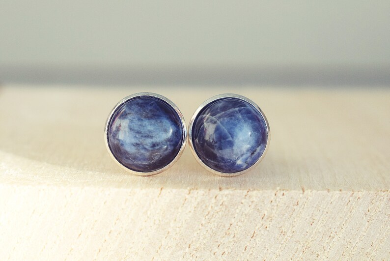May include: A pair of round stud earrings with a silver bezel. Each earring features a blue gemstone with white marbling. The earrings are displayed on a light-colored wooden surface.