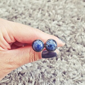 May include: A pair of round stud earrings featuring a dark blue stone with white marbling. The earrings are set in a bronze-colored metal. The earrings are held in a hand against a soft, gray background.