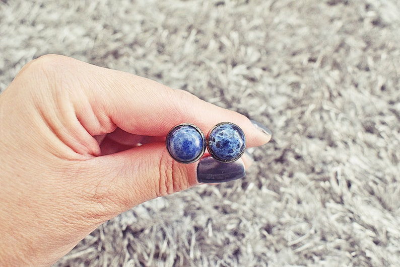 May include: A pair of round stud earrings featuring a dark blue stone with white marbling. The earrings are set in a bronze-colored metal. The earrings are held in a hand against a soft, gray background.