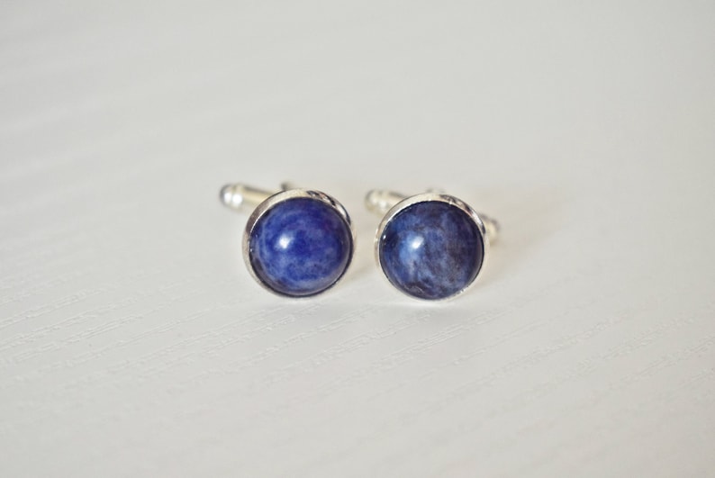 May include: A pair of cufflinks with round, deep blue stone insets. The stones have a marbled appearance and are set in a silver-colored metal. The cufflinks are displayed on a white surface.
