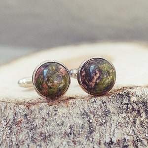 May include: A pair of cufflinks with round, polished stone faces. The stones display a mix of green and pink hues, set in silver-colored metal. The cufflinks are displayed on a textured, light brown wooden surface.