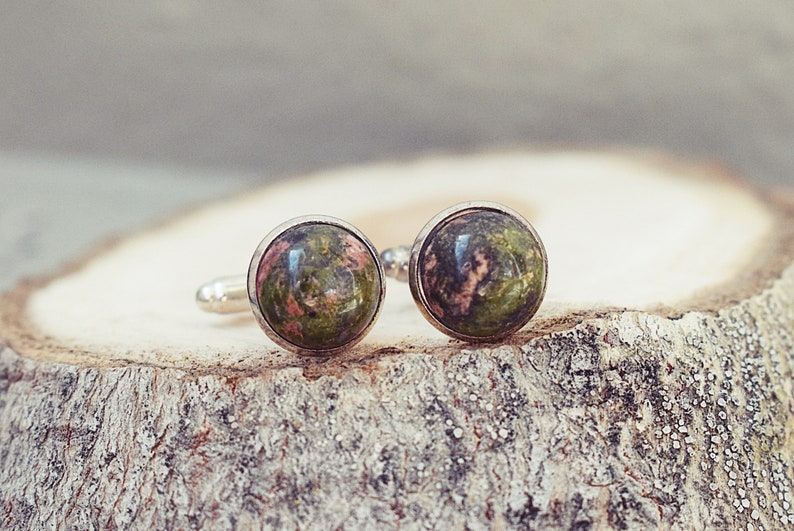 May include: A pair of cufflinks with round, polished stone faces. The stones display a mix of green and pink hues, set in silver-colored metal. The cufflinks are displayed on a textured, light brown wooden surface.