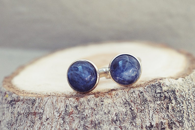 May include: A pair of silver-toned stud earrings with large, round, deep blue gemstones. The earrings are set on a natural wood slice, showcasing the jewelry's color and texture. The gemstones have a marbled appearance.