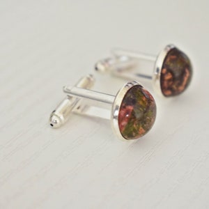 May include: A pair of silver cufflinks with teardrop-shaped, polished stone faces. The stones have a mix of red, green, and brown hues. The cufflinks are set against a white background, highlighting the details of the jewelry.