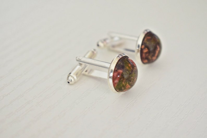 May include: A pair of silver cufflinks with teardrop-shaped, polished stone faces. The stones have a mix of red, green, and brown hues. The cufflinks are set against a white background, highlighting the details of the jewelry.