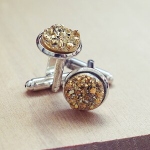 May include: A pair of silver cufflinks with round, gold-colored, crystal-like centers. The cufflinks are displayed on a light brown wooden surface. The gold centers have a textured, sparkling appearance.