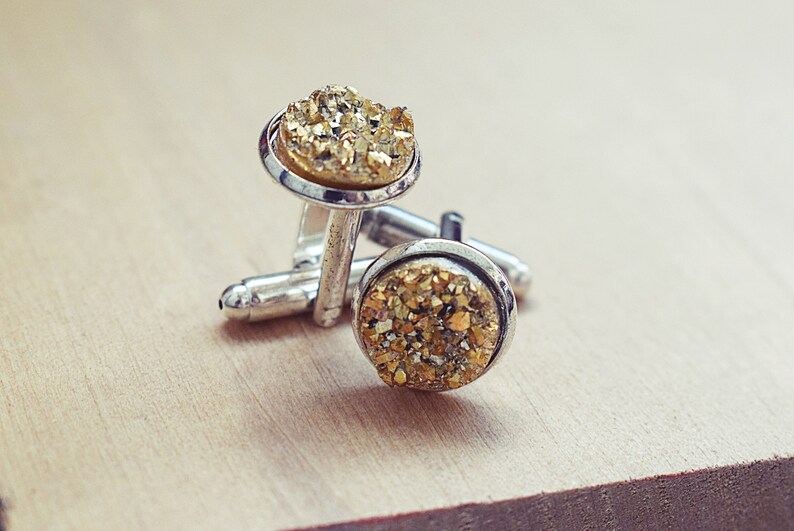 May include: A pair of silver cufflinks with round, gold-colored, crystal-like centers. The cufflinks are displayed on a light brown wooden surface. The gold centers have a textured, sparkling appearance.
