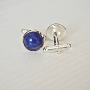 May include: A pair of silver cufflinks with a round, deep blue stone set in each cufflink. The cufflinks are displayed on a white surface, with one cufflink fastened and the other unfastened.