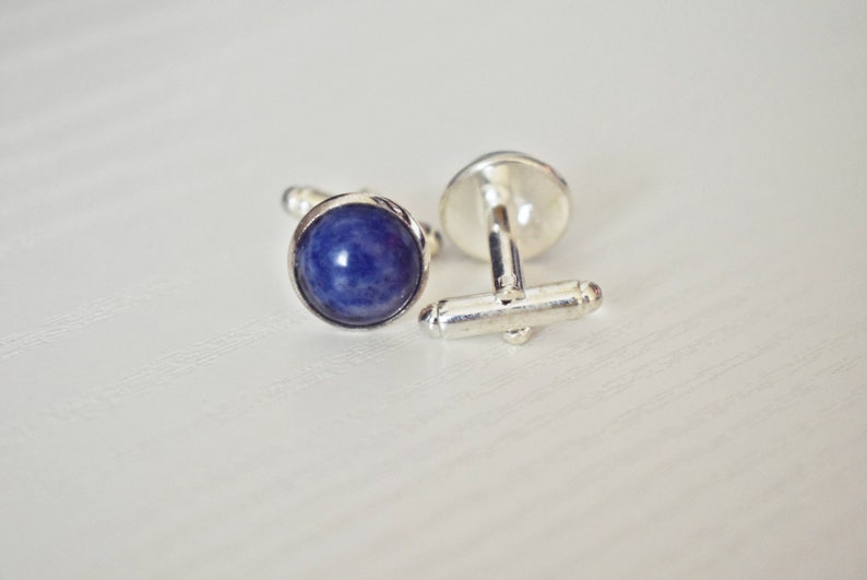 May include: A pair of silver cufflinks with a round, deep blue stone set in each cufflink. The cufflinks are displayed on a white surface, with one cufflink fastened and the other unfastened.
