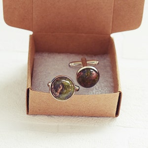 May include: A pair of cufflinks with a natural stone appearance, featuring shades of green, brown, and red. The cufflinks are presented in a small, open, brown cardboard box, resting on a white surface.