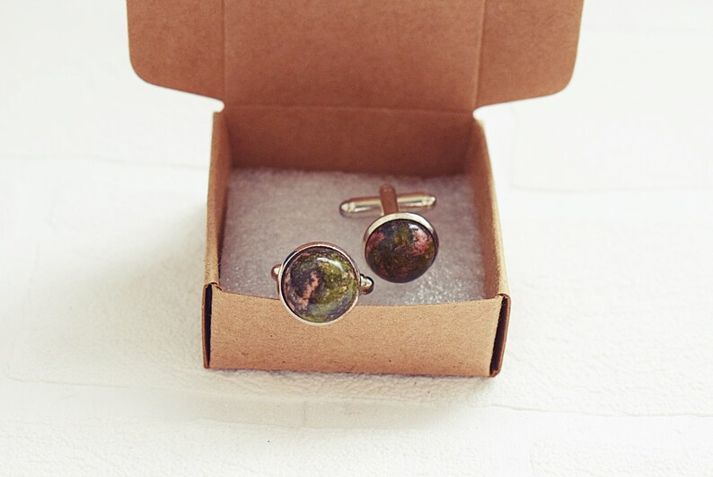 May include: A pair of cufflinks with a natural stone appearance, featuring shades of green, brown, and red. The cufflinks are presented in a small, open, brown cardboard box, resting on a white surface.
