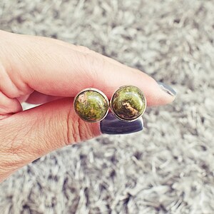 May include: A pair of round stud earrings. Each earring features a polished, green and brown stone set in a silver-colored bezel. The earrings are held between fingers against a soft, gray background.