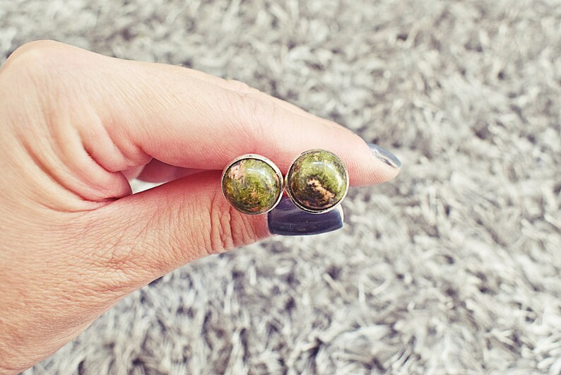 May include: A pair of round stud earrings. Each earring features a polished, green and brown stone set in a silver-colored bezel. The earrings are held between fingers against a soft, gray background.