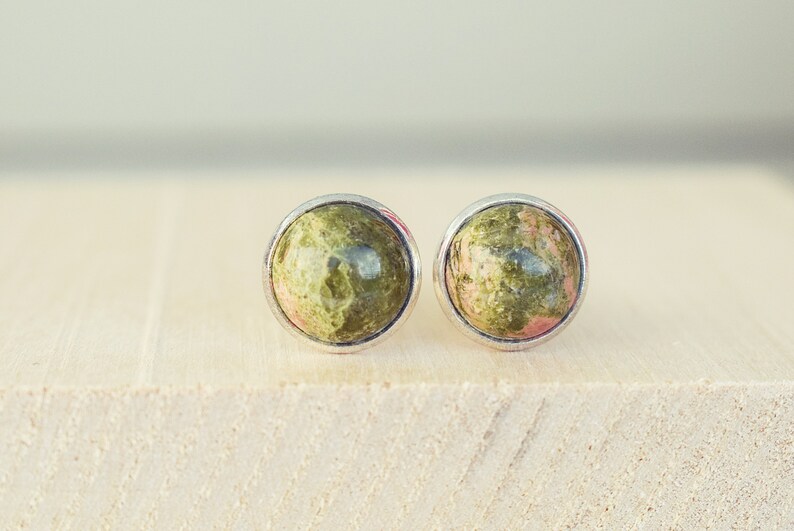 May include: A pair of round stud earrings featuring a natural stone with green and pink hues. The stones are set in a silver-colored bezel. The earrings are displayed on a light-colored wooden surface.