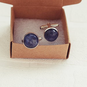 May include: A pair of round cufflinks with a silver-colored trim and a dark blue, marbled stone-like center. The cufflinks are presented in a small, open, brown cardboard box, resting on a white surface.