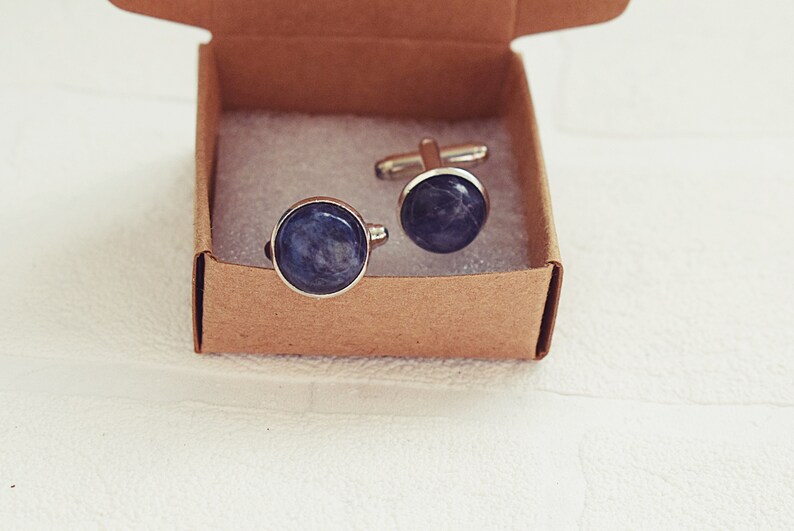 May include: A pair of round cufflinks with a silver-colored trim and a dark blue, marbled stone-like center. The cufflinks are presented in a small, open, brown cardboard box, resting on a white surface.