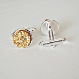 May include: A pair of silver cufflinks with round, gold-colored, textured faces. The gold faces have a sparkling, glittery appearance. The cufflinks are set against a white background.