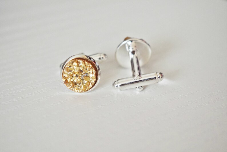 May include: A pair of silver cufflinks with round, gold-colored, textured faces. The gold faces have a sparkling, glittery appearance. The cufflinks are set against a white background.