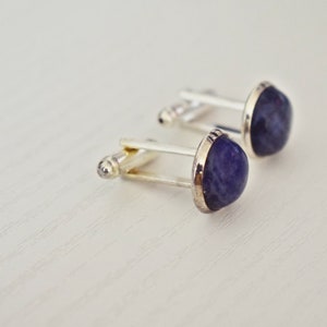 May include: A pair of cufflinks with a silver-colored metal base and a rounded, deep blue stone. The stones have a slightly irregular shape and are set within a silver bezel. The cufflinks are displayed on a light-colored surface.