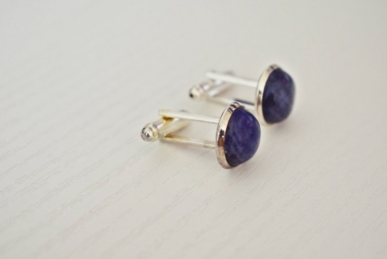 May include: A pair of cufflinks with a silver-colored metal base and a rounded, deep blue stone. The stones have a slightly irregular shape and are set within a silver bezel. The cufflinks are displayed on a light-colored surface.