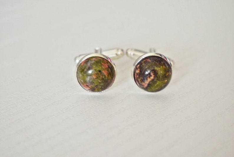 May include: A pair of cufflinks with a silver setting. Each cufflink features a round, polished stone with a mix of green, brown, and pink hues. The cufflinks are displayed on a white surface.
