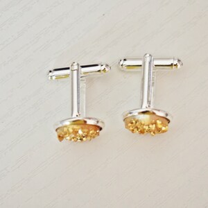 May include: A pair of silver cufflinks with a round, light-colored base and a cluster of golden-colored crystals. The cufflinks have a classic T-bar closure and are set against a white background.
