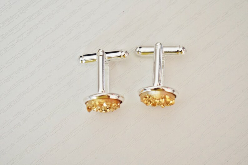 May include: A pair of silver cufflinks with a round, light-colored base and a cluster of golden-colored crystals. The cufflinks have a classic T-bar closure and are set against a white background.