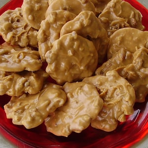 May include: A close-up shot of a plate filled with peanut brittle candies. The candies are a light brown color with a glossy finish and a textured surface. They are arranged on a red plate with a silver rim.