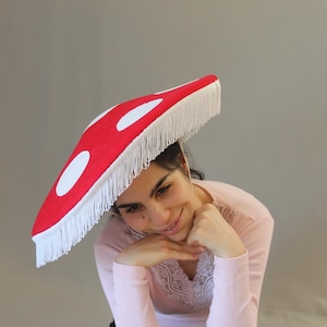 Può includere: Un cappello a tesa larga a forma di cappello di fungo, caratterizzato da una parte superiore rosso vivace con grandi macchie circolari bianche. Il bordo del cappello è rifinito con una lunga frangia bianca che pende lungo il perimetro.