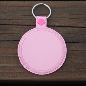 May include: A pink, round keychain with a silver key ring. The keychain has a stitched border and a small pink flower accent. The background is a dark wood surface.