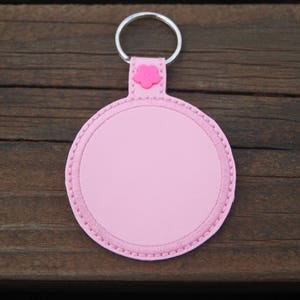 May include: A pink, circular keyring with a silver ring. The keyring features a stitched edge and a small flower detail. Set against a dark wooden background.