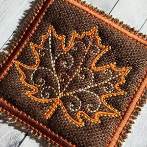 May include: A brown burlap square with an orange and white embroidered maple leaf design. The square has a brown and orange fringe border.