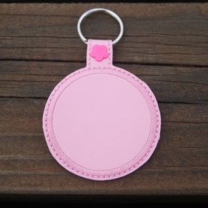 May include: A pink, round keychain with a silver key ring. The keychain has a stitched border and a small pink flower accent. The background is a dark wood surface.