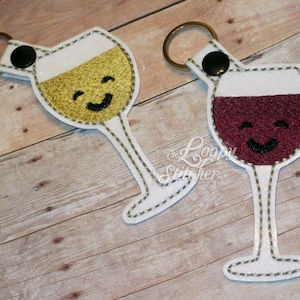 May include: Two white wine glass keychains with key rings. One glass is filled with a yellow liquid and the other is filled with a red liquid. Both glasses have a smiling face embroidered on them.